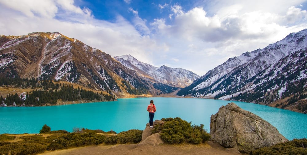 A woman is standing on top of a mountain overlooking a blue lake in Canada.