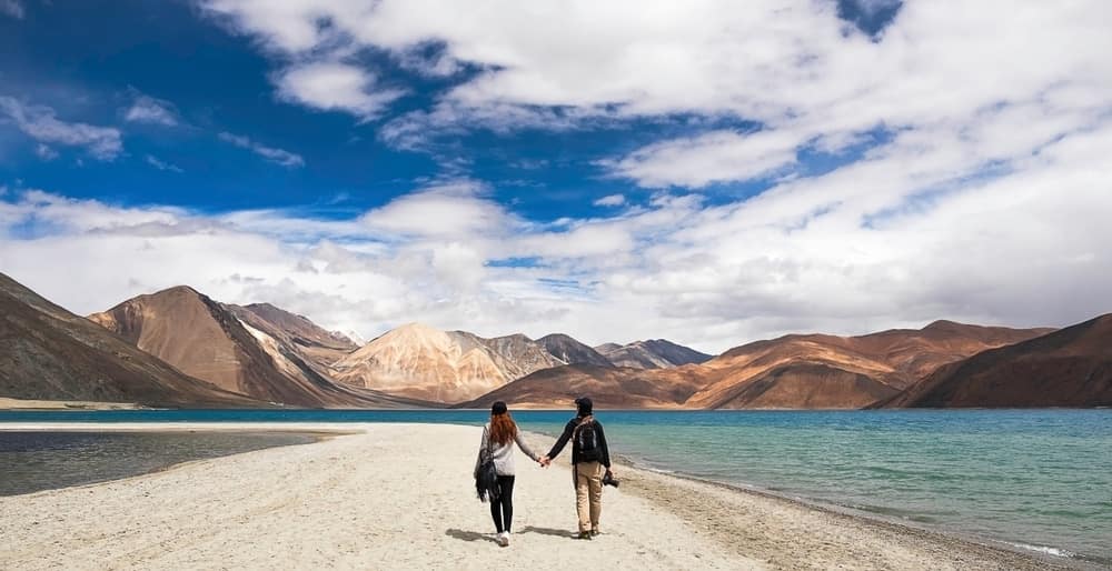 A couple enjoying Caravan Tourism on India's Coastal Districts, strolling along a picturesque beach with majestic mountains in the background.