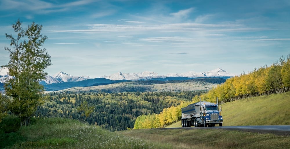 A Canadian semi truck driving down a country road with mountains in the background.