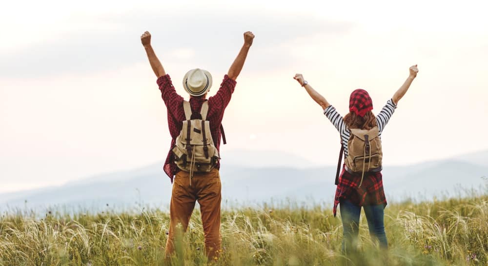 Two people with backpacks standing in a field with their arms raised.