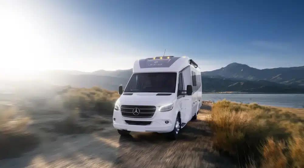 The Mercedes-Benz RV, with its Unity Corner Bed and RV Luxury features, is cruising down a dirt road near a lake.
