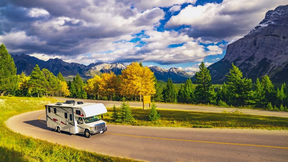 An RV with units travels down a winding road with mountains in the background.