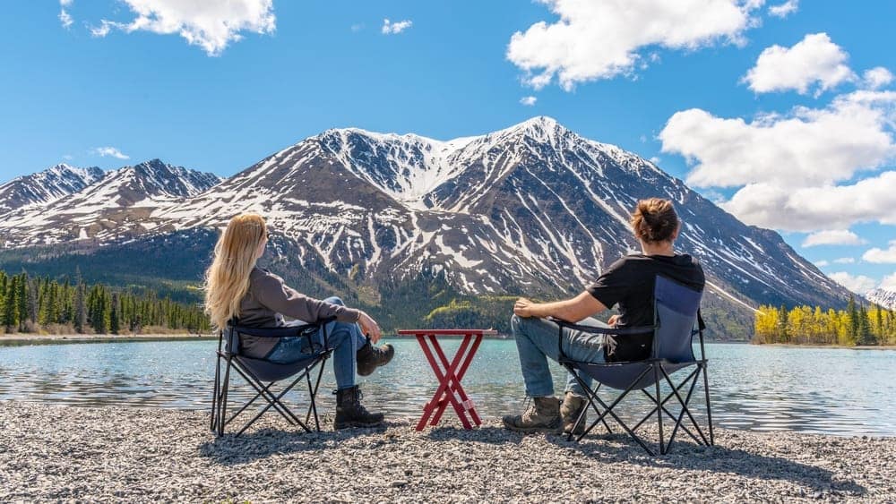 Two people from Indigenous groups sitting in chairs on the shore of a lake with mountains in the background.