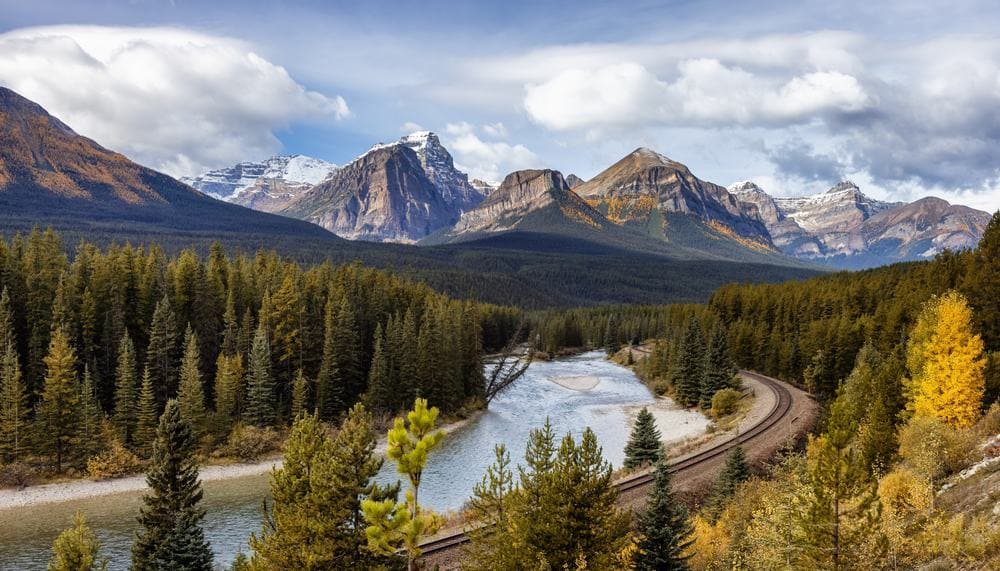 A train traveling along a river with breathtaking mountains in the background.