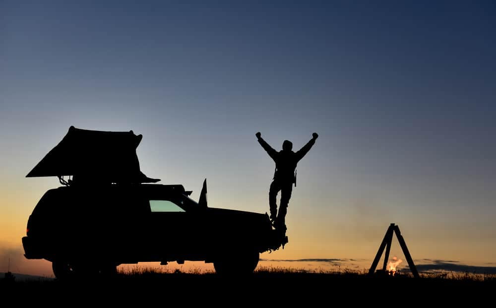 Silhouette of a man standing on top of a car's roof at sunset, next to an Easy-To-Set-Up Tent.