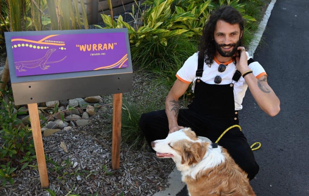 A man with a dog sitting next to a sign promoting the Aboriginal Art Trail in NSW, showcasing the rich culture.