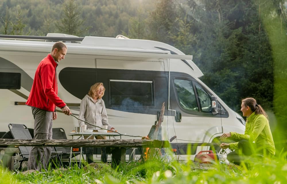 New members of an RV travel group gather around a campfire in front of an RV, enjoying the scenic Idaho wilderness.