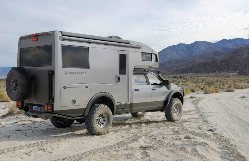 A silver truck with a camper, an Expedition Vehicle, parked on the side of a dirt road.