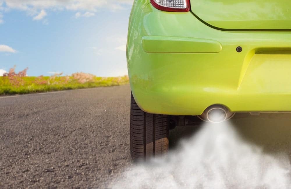 A green car is driving down the road with smoke coming out of its tailpipe, highlighting the need for stricter fuel economy regulations under the Biden Administration.