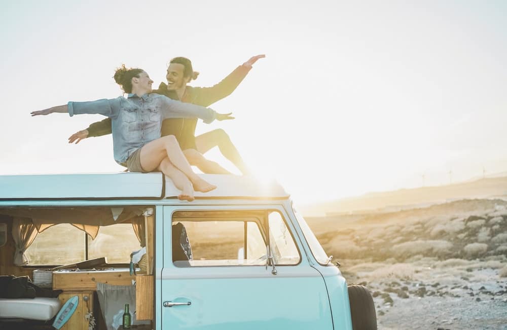 Stylish travelers sitting on top of a van with their arms outstretched at an RV park.