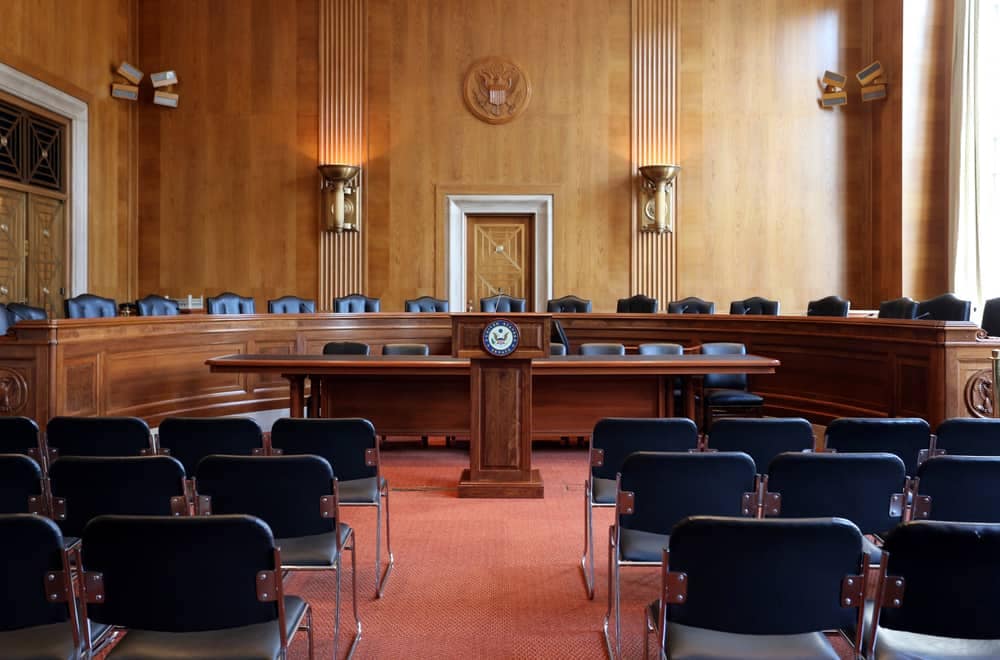 An empty courtroom with black chairs, a wooden table, and plan approval delays.