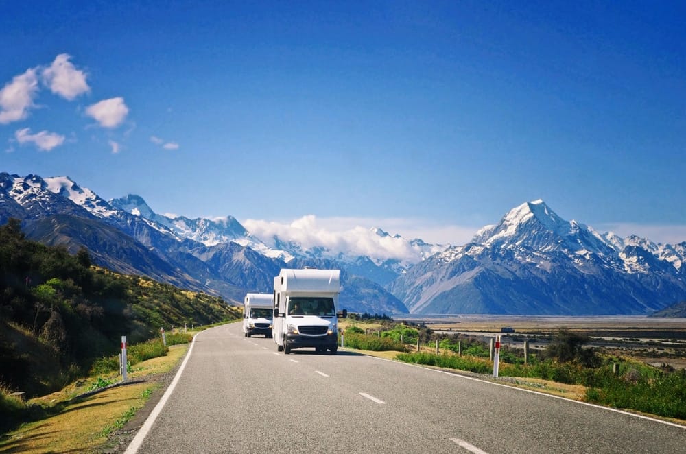 Two rvs driving down a road with mountains in the background.