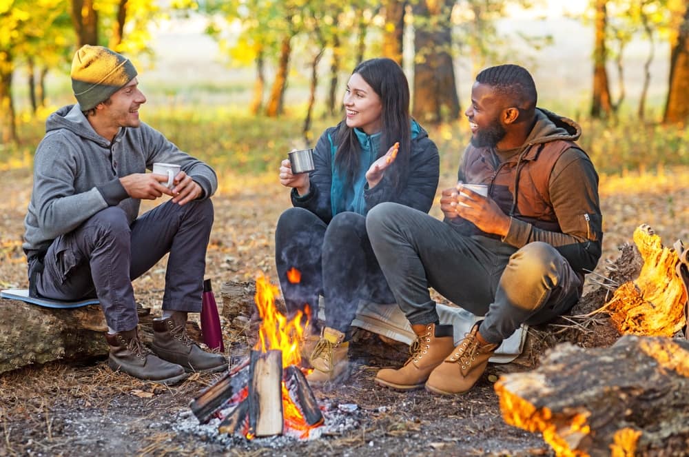 Three people sitting around a campfire at a campground in the woods.
