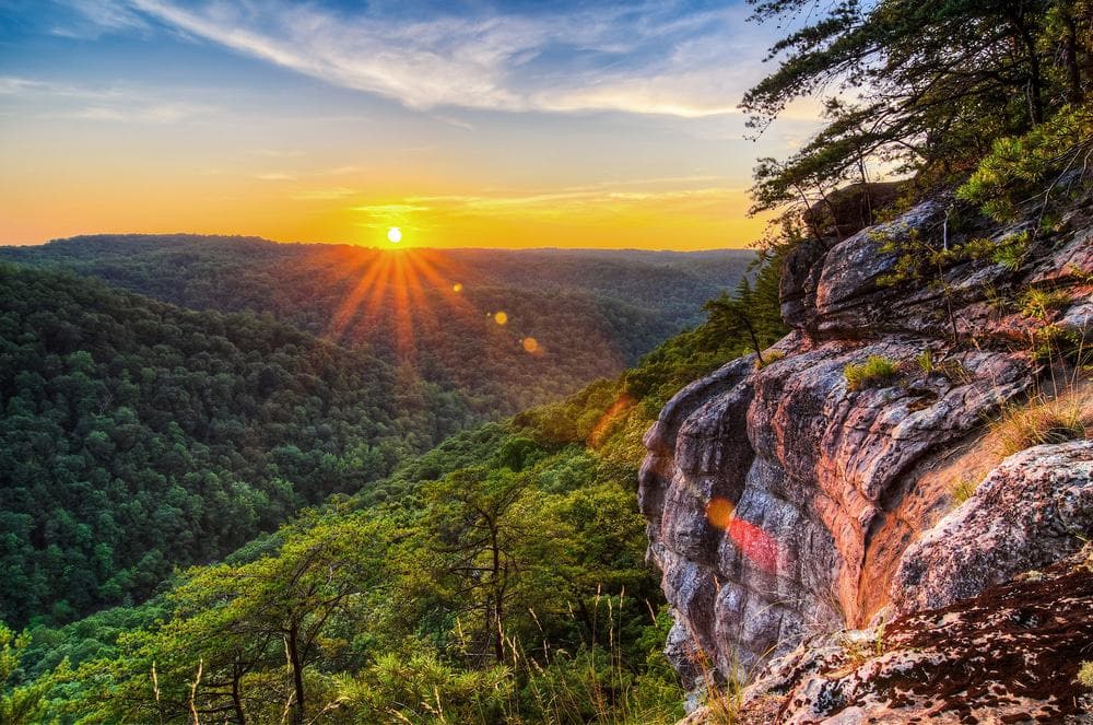 The sun rises over a rocky cliff overlooking a forest.