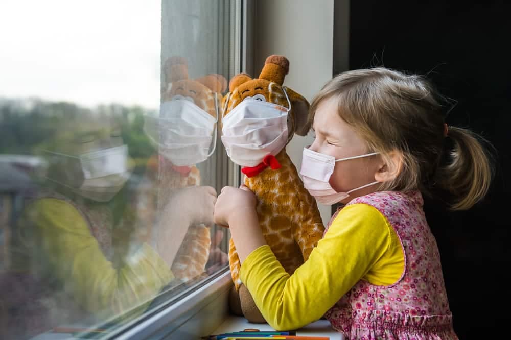 A little girl wearing a **surgical mask** and holding a stuffed giraffe.