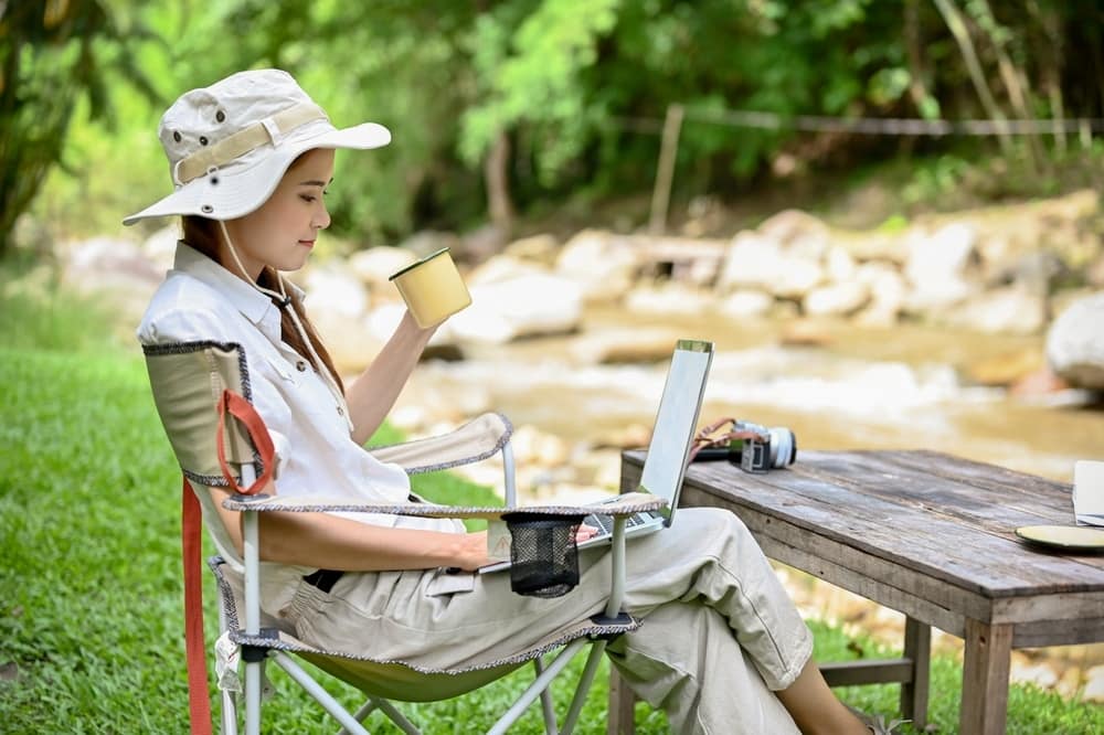 A woman in a hat is sitting in a chair and using a laptop to browse through significant achievements on her 1,700th virtual tour of campground views.