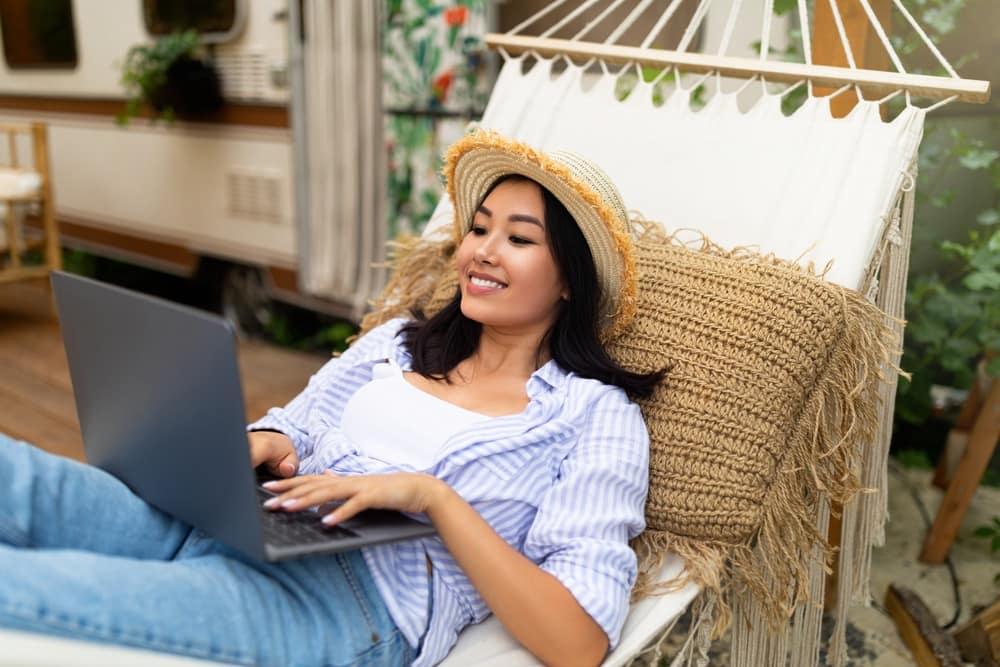 A woman in a hat is sitting in a hammock with a laptop.
