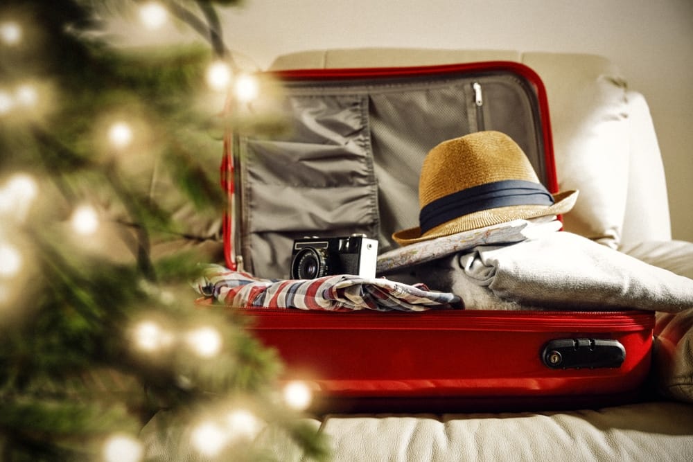 A red suitcase with clothes and a hat in front of a Christmas tree, perfect for gifting.