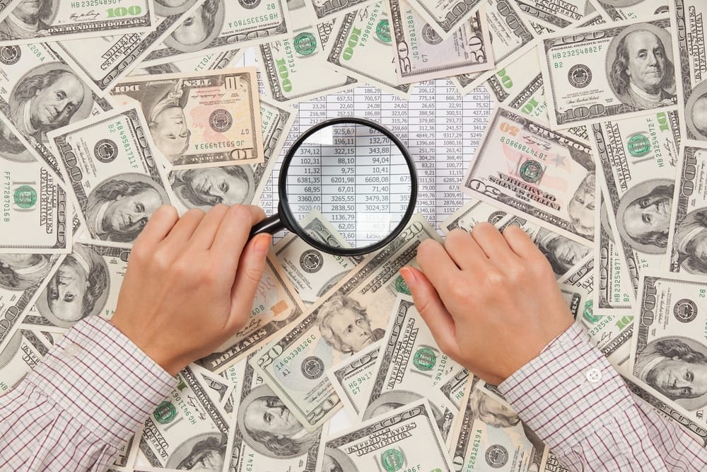 The Fed pulls back on economic aid, as hands hold a magnifying glass over a pile of money.