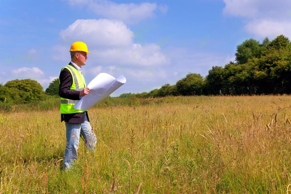 A man in a hard hat standing in a field with blueprints.