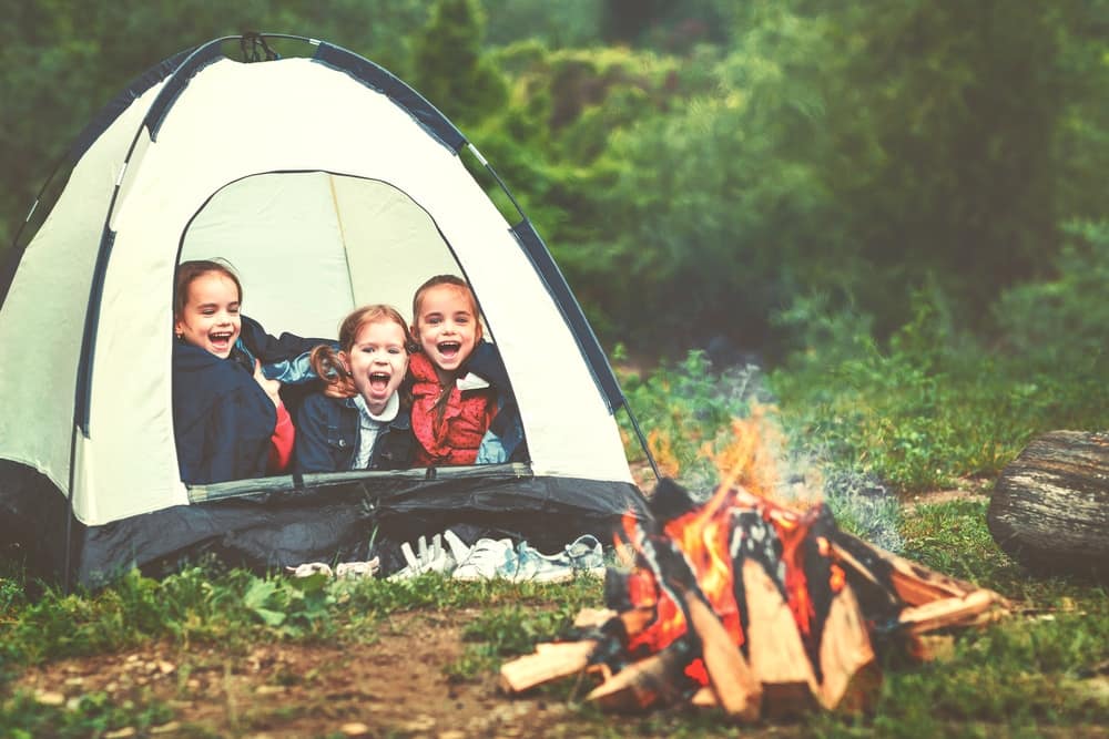 Three children sitting in front of an RV near a campfire.