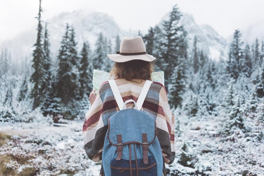 A woman wearing a hat and backpack is standing in a snowy forest, perfect for U.S. travelers seeking winter trips.