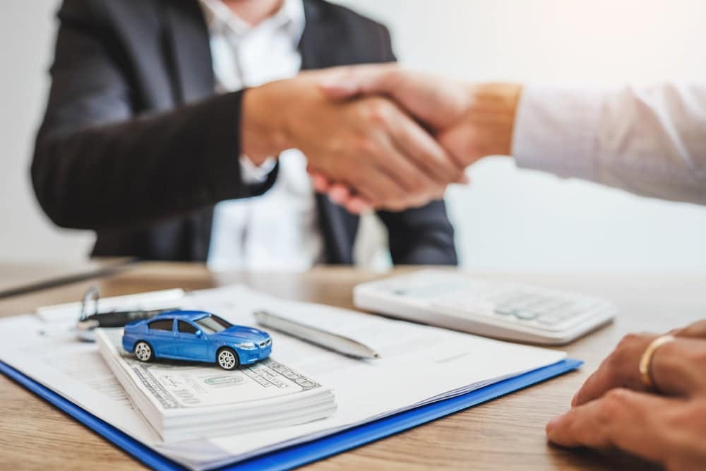 A man shaking hands with a blue car, sold next year, on a desk.