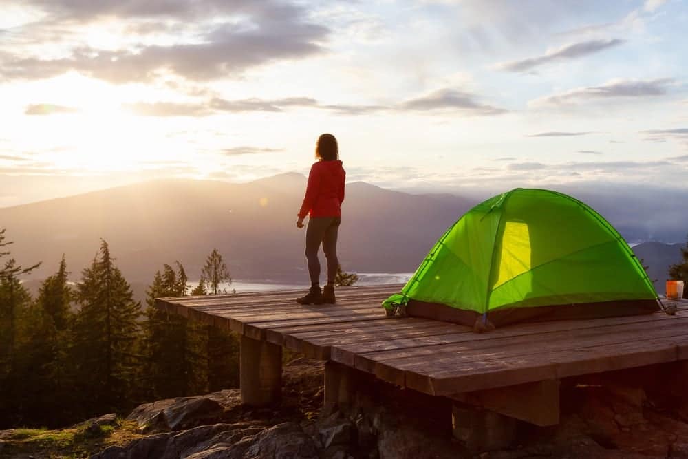 A woman standing next to a green tent at sunset, enjoying a moment of commercial recreation.