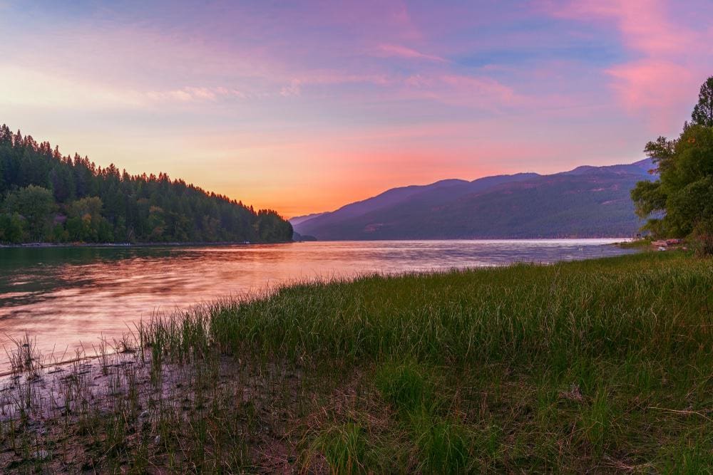 A picturesque sunset over Flathead Lake with grassy shores and majestic mountains in the background, perfect for a visitation to Flathead Lake State Parks.