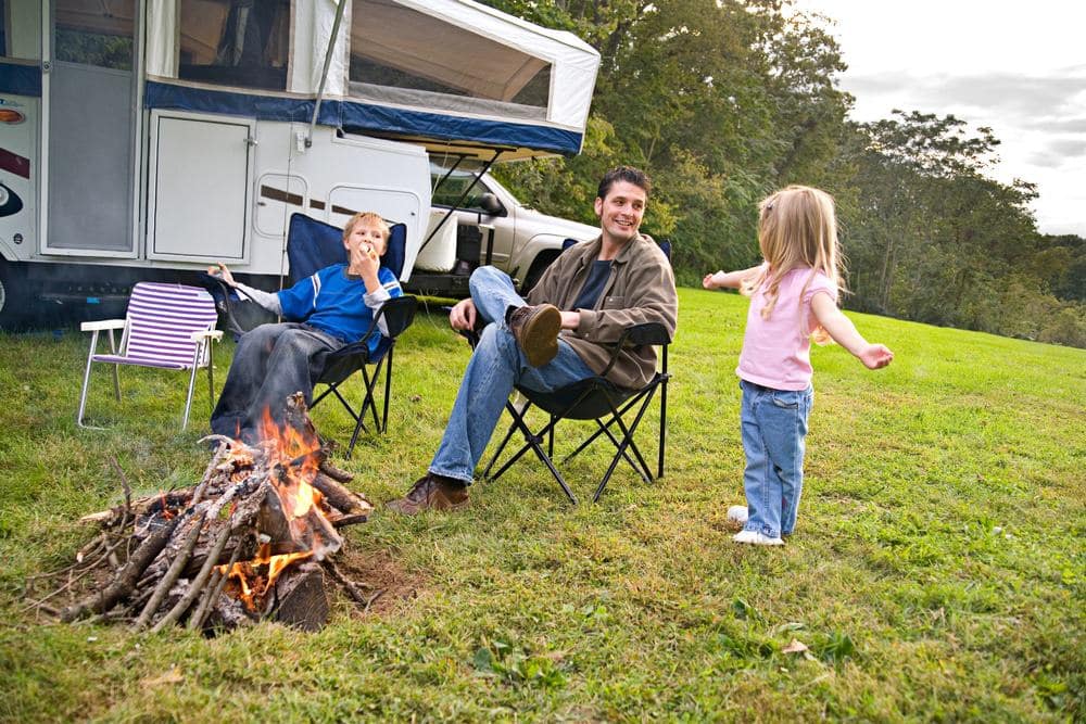 A family sits around a campfire in front of an RV.