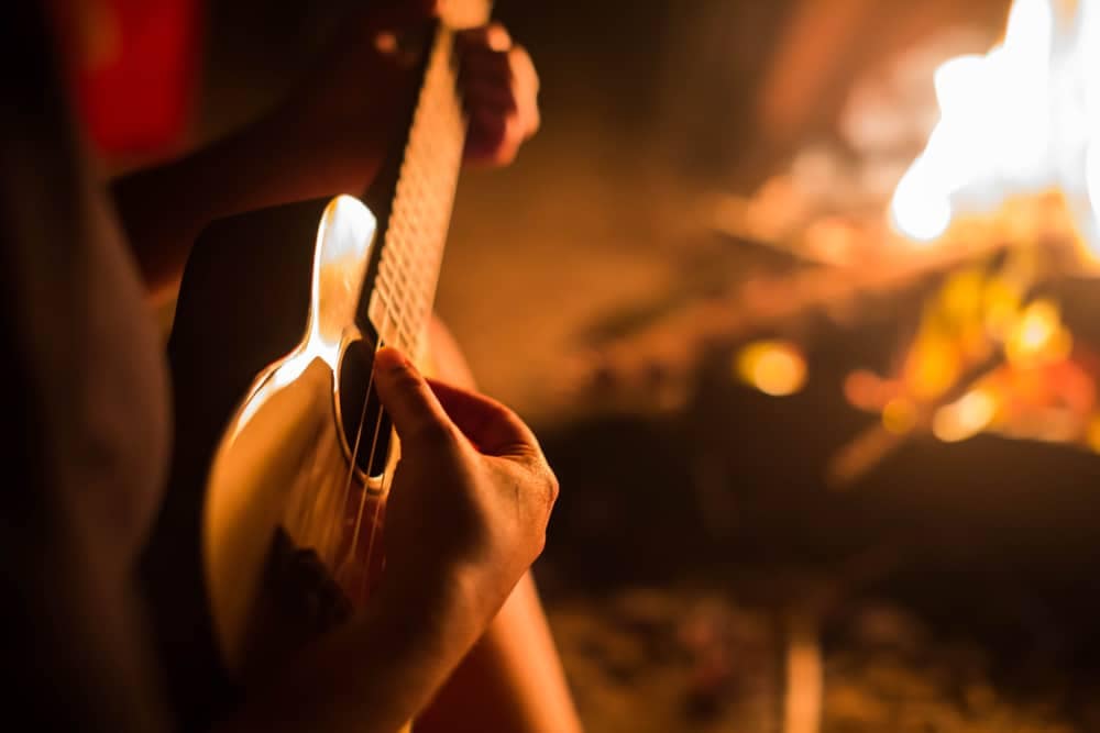 A person playing an ukulele in front of a fire at Holiday Park.