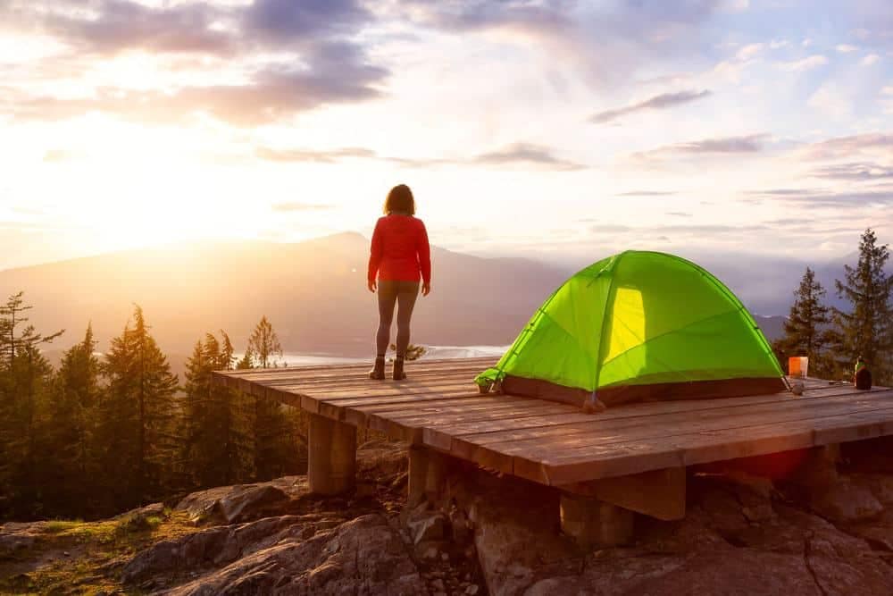 A woman is standing next to a tent in the mountains at sunset.