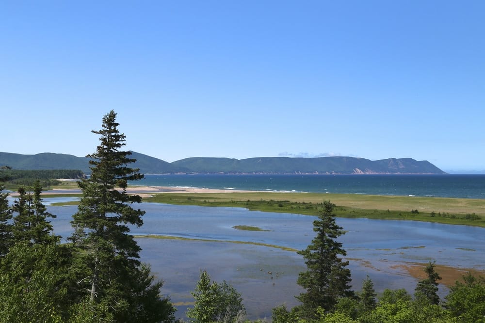 A serene view of a lake surrounded by lush trees and majestic mountains in the distance at Cape Breton, perfect for escaping the busy season at campgrounds.