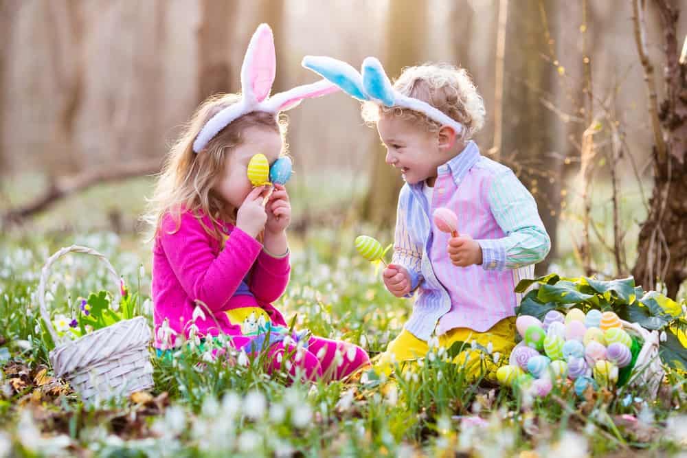 Two children excitedly participate in an Easter Egg Scavenger Hunt at Camp Helen State Park, wearing bunny ears and surrounded by a picturesque woodland setting.