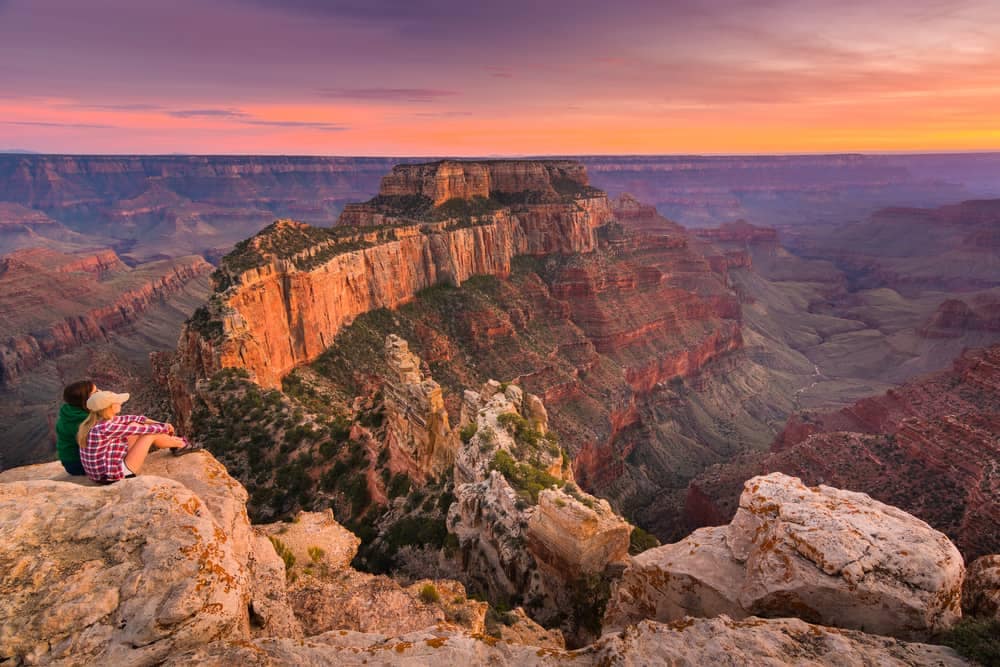 Two people sitting on the North Rim of the Grand Canyon at sunset.