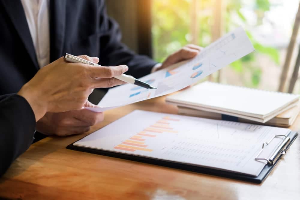 A businessman in a suit is holding a pen and looking at a piece of paper displaying Q1 Revenue.