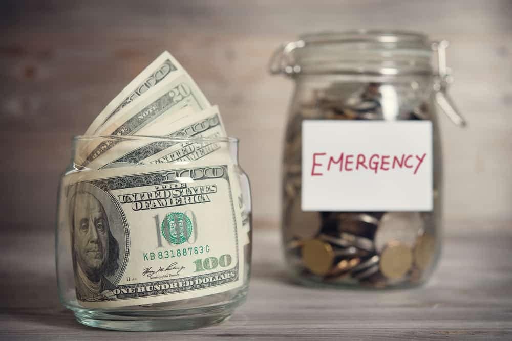 A glass jar filled with money next to a sign that says Emergency Funding.