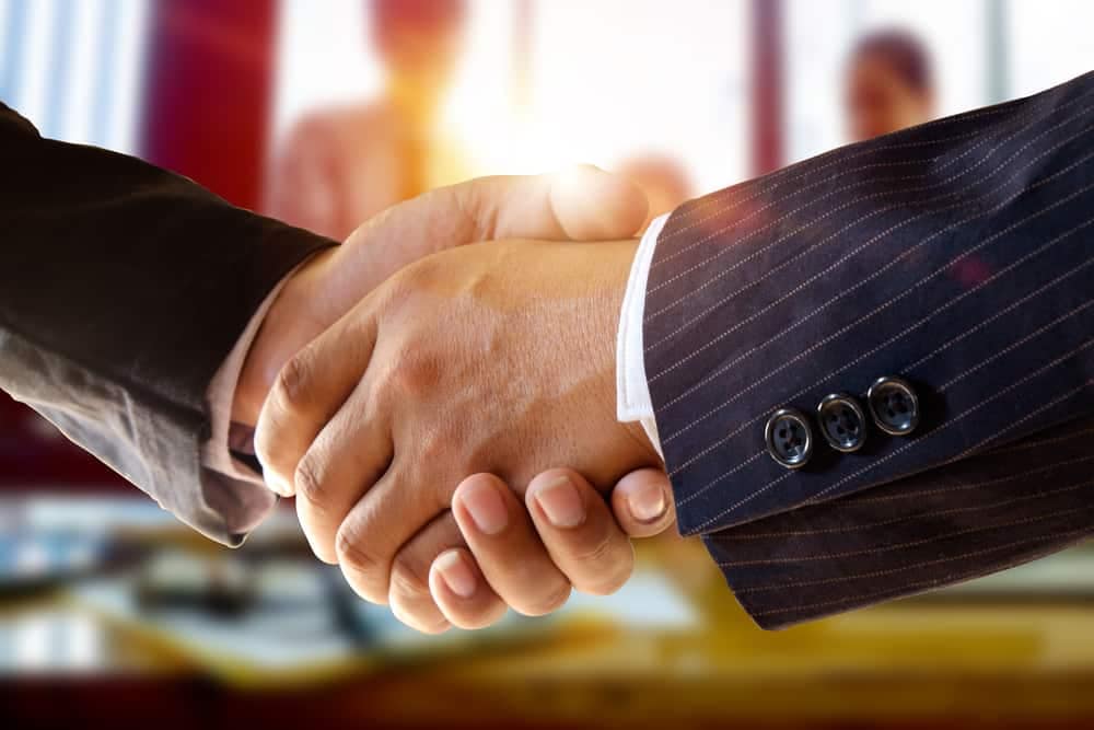 Two business people shaking hands in front of a group of people at Acquires.