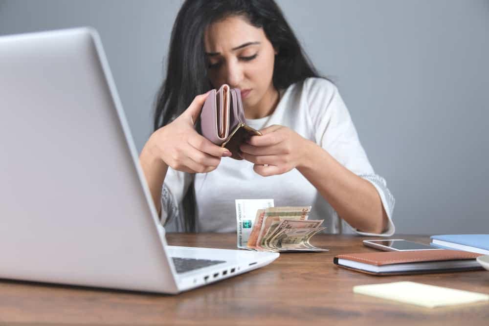 A woman, concerned about consumer confidence, slides down in her chair while looking at her wallet on a desk with a laptop.