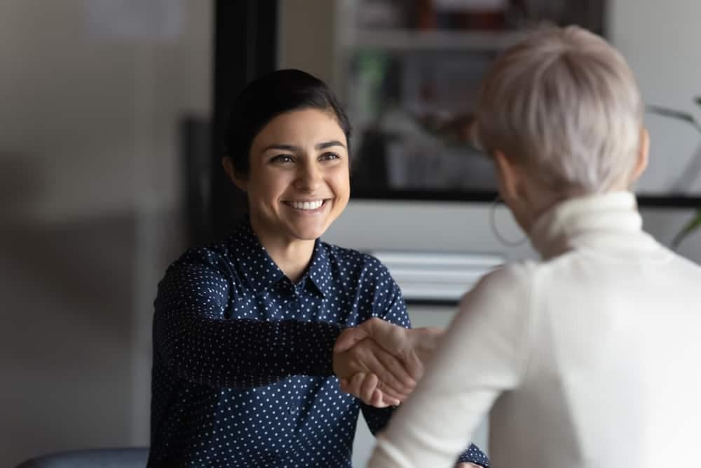 A woman shakes hands with another woman in an office during a meeting discussing Board Of Director Positions.