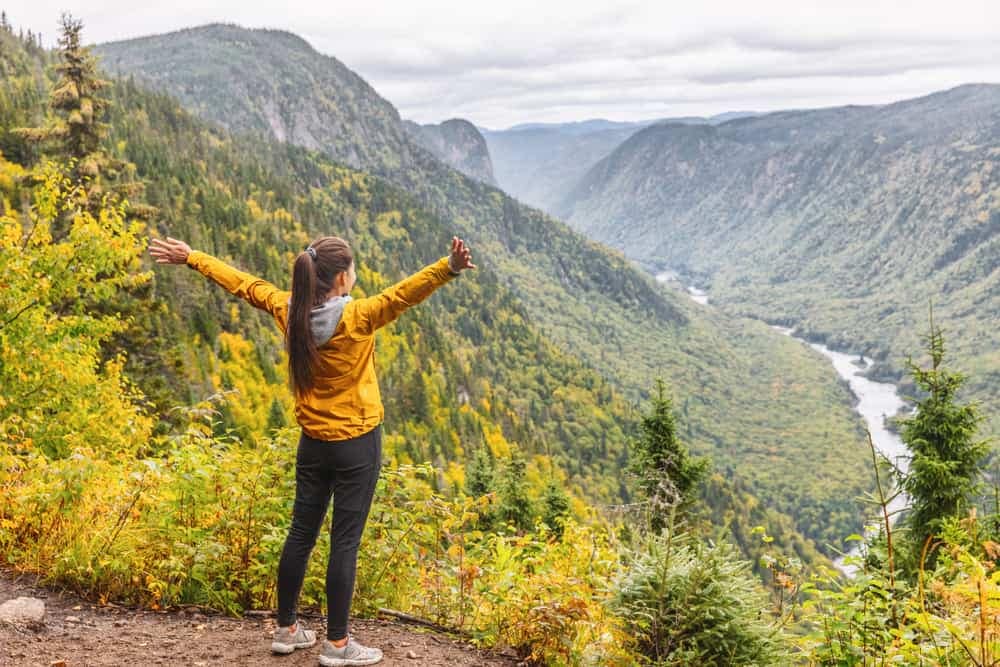 A woman with her arms outstretched is standing on a mountain overlooking a river, feeling grateful for the stunning view and the serene campgrounds nearby.