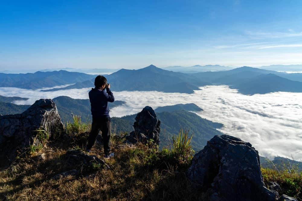 A woman is standing on top of a mountain, having the perfect holiday experience while enjoying the breathtaking view of clouds.