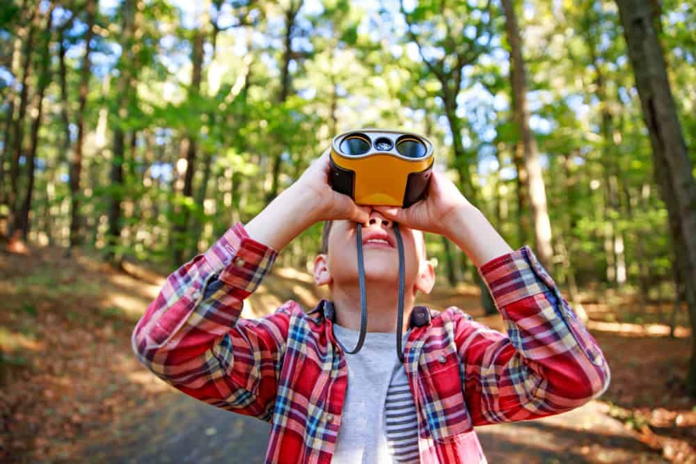 A boy on a Great Outdoor Adventure looking through binoculars in the woods.