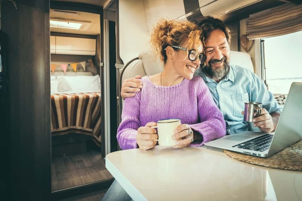A couple sitting in an RV with a laptop and a cup of coffee, making reservations using ReserveAmerica.com.