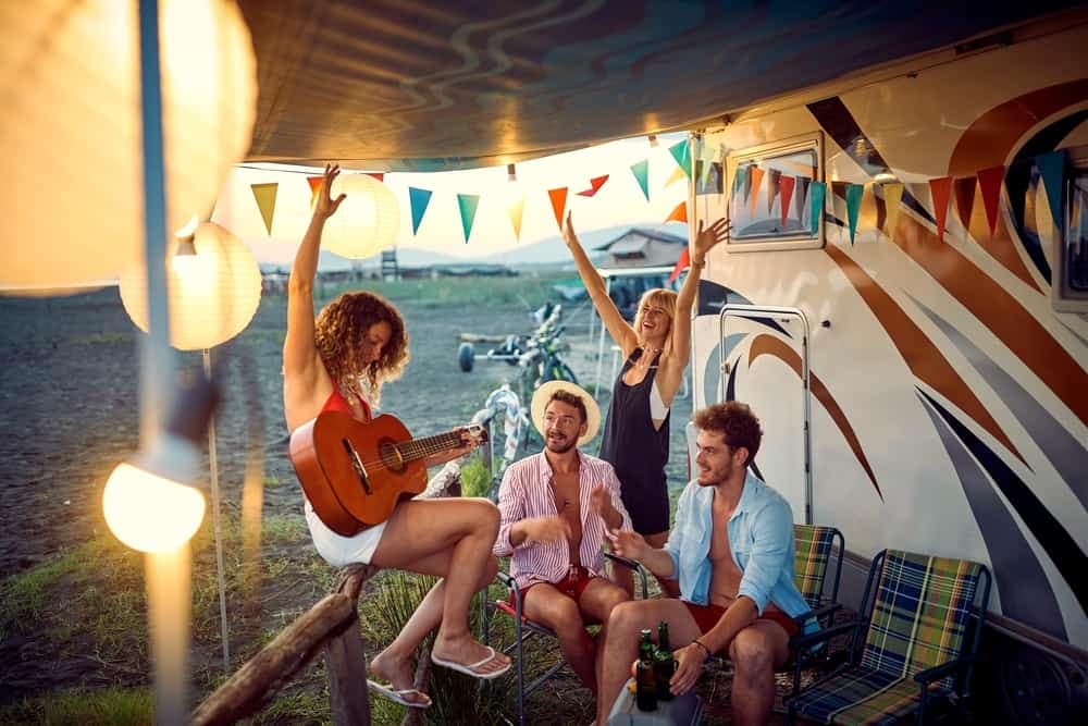 A group of campers sitting on the back of an RV, playing guitar in a park.