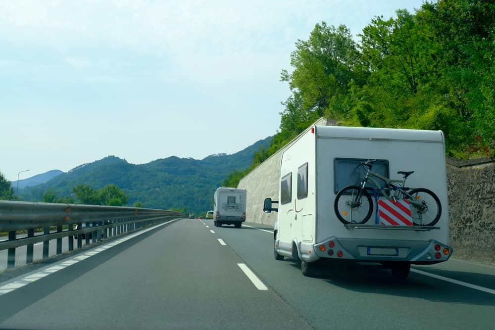 A mobile app designed for RV travelers that helps in planning trips and routes, with the added convenience of an RV equipped with a bicycle rack driving down a scenic highway.