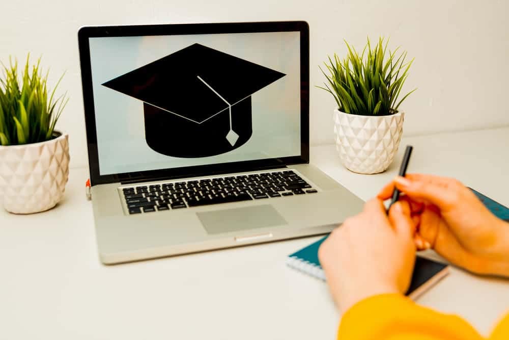 A person writing on a laptop with a graduation cap on it, applying for the Mike Molino RV Learning Center College Scholarship Program.