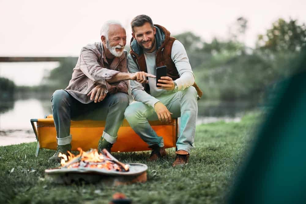 Two men sitting around a campfire in Manitoba with a cell phone, discussing the launch of a reservation system.