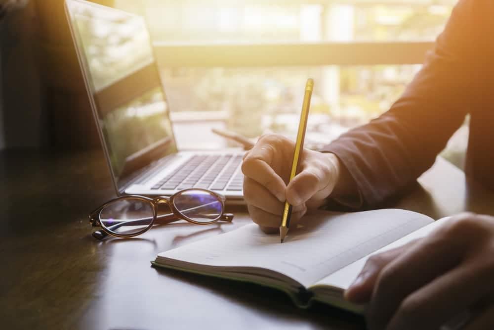 A businessman taking notes on a notebook while working on his laptop, possibly researching prospective owners for a mastery course in an RV destination.
