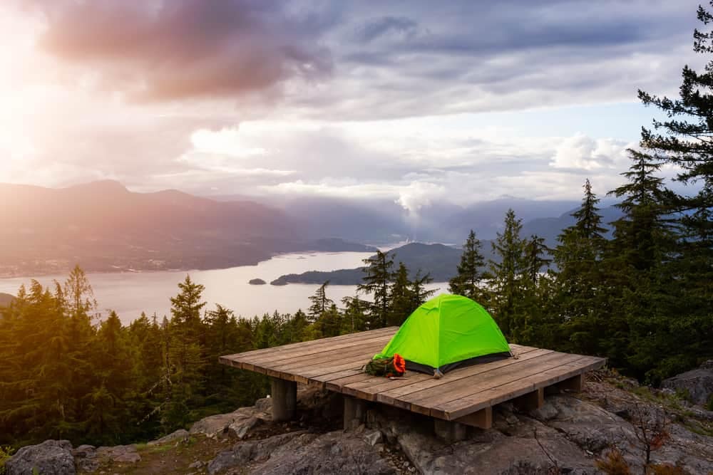 A green tent sits on top of a mountain overlooking a waterfront park.